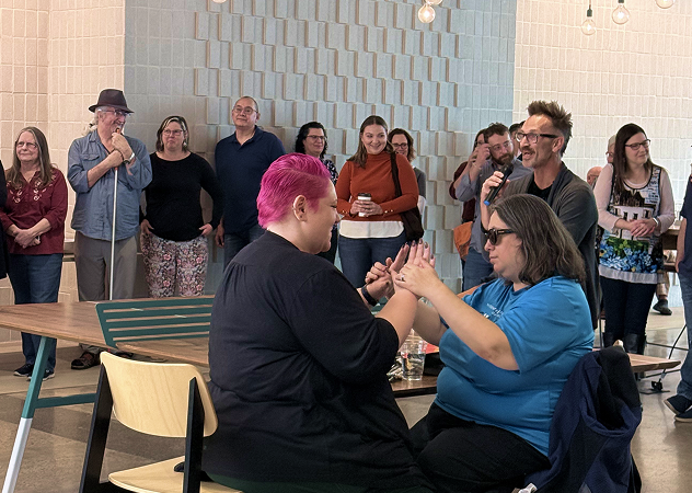 Group of people gathered indoors as two individuals communicate using sign language, highlighting accessibility and inclusion.