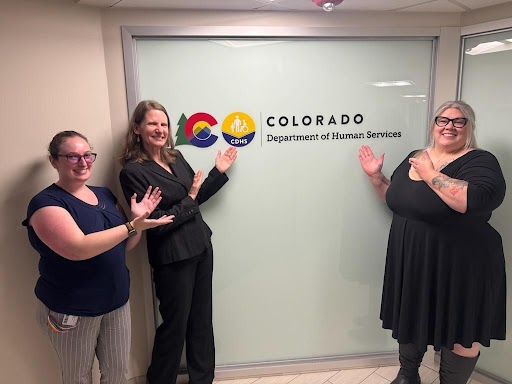 Few of Colorado Division for the Deaf, Hard of Hearing, and DeafBlind  team members standing together in an indoor hallway with Colorado Department of Human Services logo on office window panels.